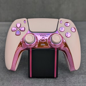 Könnte beinhalten: Ein kabelloser Videospiel-Controller mit einer matten beigen Schale und einem metallisch-rosa verchromten Mittelteil. Der Controller verfügt über passende metallisch-rosa Tasten, zwei beige Analogsticks und eine schwarze Ladestation mit rosa Streifen.