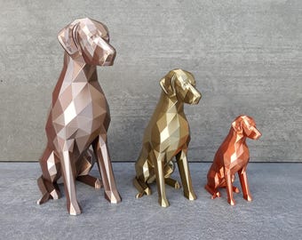 May include: Three geometric dog figurines in descending order of size. The largest is a metallic rose gold, the middle is a metallic gold, and the smallest is a metallic copper. Each dog is depicted in a seated position with faceted surfaces.