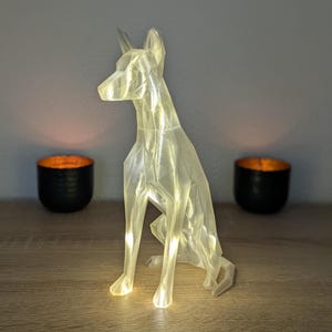 Può includere: Una statuetta di cane geometrica, traslucida e color bianco sporco, realizzata con un design sfaccettato a basso numero di poligoni. La scultura è illuminata internamente, emettendo un bagliore caldo che ne esalta i bordi angolari.