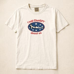 Könnte beinhalten: Cremefarbenes T-Shirt mit dem Aufdruck "I told ChatGPT about us". Die Grafik zeigt eine Frau in einem schwarzen Badeanzug in einem blauen Oval mit weißen Sternen. Das Shirt hat einen Rundhalsausschnitt und kurze Ärmel.