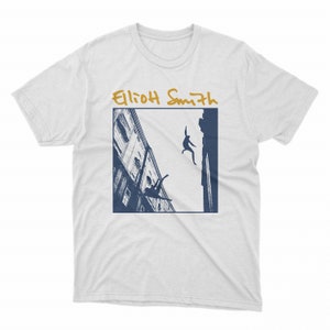 May include: White t-shirt with a blue and white image of two people falling from a building. The text "Elliot Smith" is written in gold above the image.