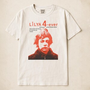 May include: Cream-colored t-shirt with a red graphic featuring the text "Lilya 4-ever" and a portrait of a person. The shirt also includes text in French and Italian, and the Comfort Colors logo.