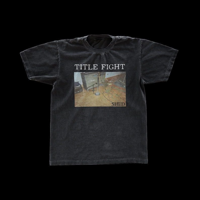 Puede incluir: Camiseta negra con un aspecto descolorido que presenta un logotipo de banda para "TITLE FIGHT" con el texto "SHED" debajo del logotipo. El logotipo es una foto de una habitaci&oacute;n con un soporte de micr&oacute;fono y un amplificador de guitarra.