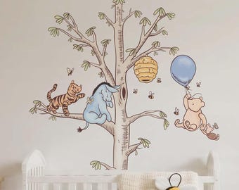 May include: A wall decal featuring a large tree with Tigger, Eeyore, and Winnie the Pooh holding a blue balloon. A yellow beehive hangs from a branch, and small bees are scattered around. The decal is positioned above a white wooden crib.