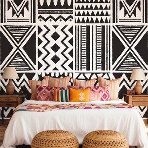Könnte beinhalten: Ein weißes Bett mit einem weißen Bettbezug und einer Vielzahl von bunten Kissen. Das Bett steht vor einer Wand mit einem schwarz-weißen geometrischen Muster. Vor dem Bett stehen zwei gewebte Hocker.