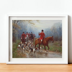 May include: Framed print of a hunting scene. A group of riders in red coats on horseback lead a pack of hounds along a wet road. The artwork is set in a forest with a soft, muted color palette, and is framed in a white border.