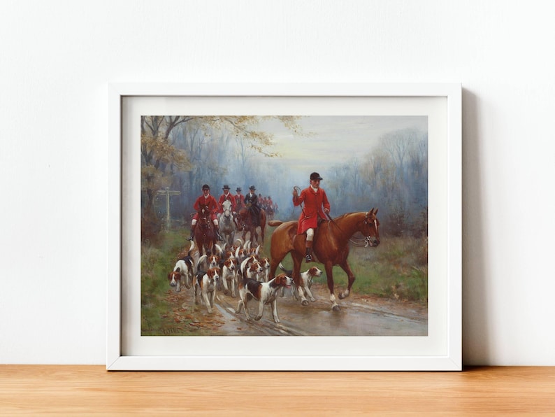 May include: Framed print of a hunting scene. A group of riders in red coats on horseback lead a pack of hounds along a wet road. The artwork is set in a forest with a soft, muted color palette, and is framed in a white border.