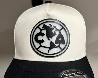 Op de afbeelding: Witte en zwarte baseballpet met een zwart-wit logo van een voetbal met een wereldkaart erin. Het logo zegt "CA" bovenaan en "SNAPBACK" onderaan.