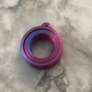 May include: A circular, 3D-printed object featuring a gradient of purple, blue, and magenta colors. The item consists of two concentric rings with a small loop attached to the outer edge, displaying visible horizontal layer lines from the printing process.