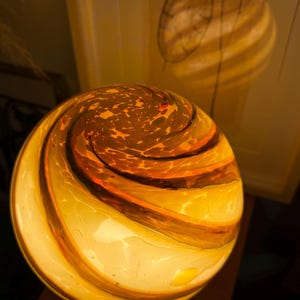 May include: A glowing, spherical glass lamp with a swirling pattern of amber, orange, and white. The lamp casts a warm light, creating a cozy ambiance. The design features a textured surface and a unique, artistic aesthetic.