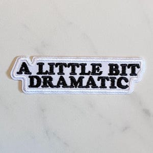 May include: A white patch with black text that reads "A LITTLE BIT DRAMATIC." The patch has a black border and is rectangular in shape. The patch is on a white, hexagonal surface.