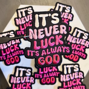 May include: Embroidered patches with the text "IT'S NEVER LUCK IT'S ALWAYS GOD" in white, pink, and red. The patches are black with a white border and are arranged on a white hexagonal surface. A pair of tweezers is holding one patch.