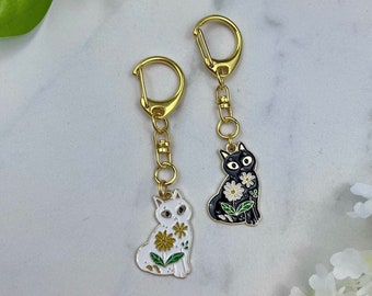 May include: Two cat-shaped keychains. One is white with yellow and green floral accents, the other is black with white daisy accents. Both have gold-colored hardware and a clasp for attaching to keys or bags. The keychains are set against a white and gray background.