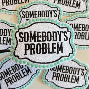 May include: A collection of white embroidered patches with the text "SOMEBODY'S PROBLEM" in black. Each patch has a decorative border with a light green trim and a black outline. The patches are arranged on a light-colored surface.