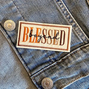 May include: A rectangular patch with the words "BLESSED beyond" embroidered in orange, brown, and black thread. The patch is bordered in brown and is affixed to a light blue denim jacket.