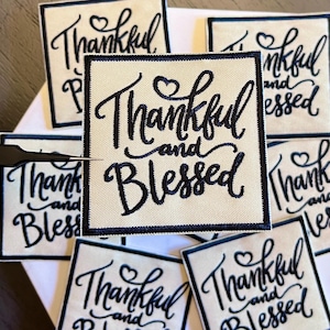May include: A collection of square patches with the words "Thankful and Blessed" in elegant black script. Each patch has a cream-colored background and a black border. A pair of tweezers is visible.