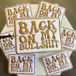 May include: Embroidered patches with the phrase "BACK ON MY BULLSHIT" in gold thread on a white background. The patches are square with a gold border and are arranged on a hexagonal surface.