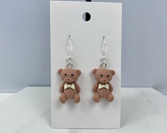Può includere: Un paio di orecchini pendenti a forma di orsacchiotto marrone con un papillon bianco. Gli orecchini sono su una carta bianca.