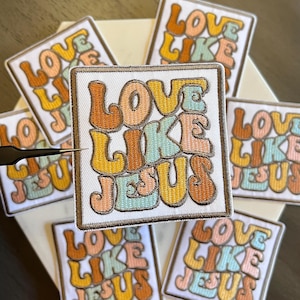 May include: Embroidered patches with the text "LOVE LIKE JESUS" in a retro font. The patches feature a white background with the text in shades of orange, yellow, and teal. The patches are square and have a brown border.