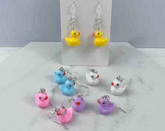 May include: A set of colorful rubber ducky earrings. The earrings are silver and feature a variety of colors including yellow, blue, white, pink, and purple.