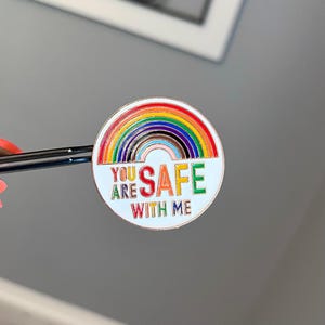 Pu&ograve; includere: Spilla rotonda in smalto con un design arcobaleno e la scritta "YOU ARE SAFE WITH ME" in lettere colorate. La spilla ha uno sfondo bianco e un bordo argentato. La spilla &egrave; tenuta da una pinzetta.