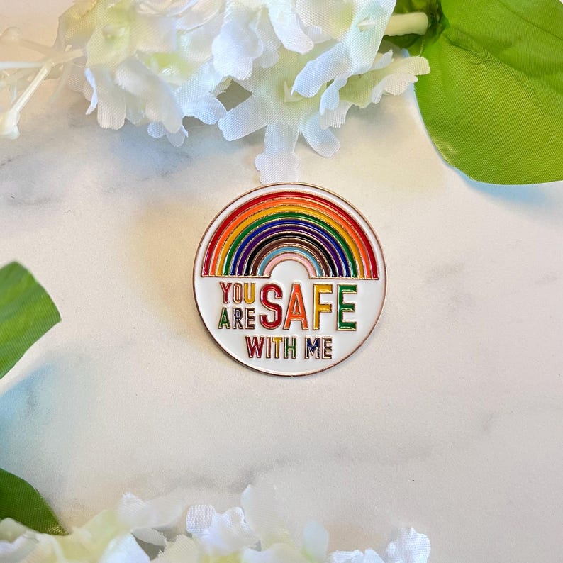 Pu&ograve; includere: Spilla rotonda in smalto con un design arcobaleno e le parole "YOU ARE SAFE WITH ME" in lettere colorate. La spilla ha un bordo bianco ed &egrave; posta su uno sfondo bianco con accenti floreali.
