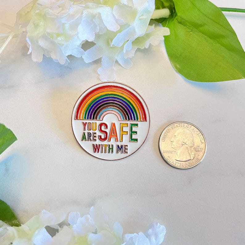 Pu&ograve; includere: Spilla rotonda in smalto con un design arcobaleno e le parole "YOU ARE SAFE WITH ME" in bianco, verde e rosso. Una moneta &egrave; accanto alla spilla. La spilla &egrave; su una superficie bianca con fiori bianchi e foglie verdi.