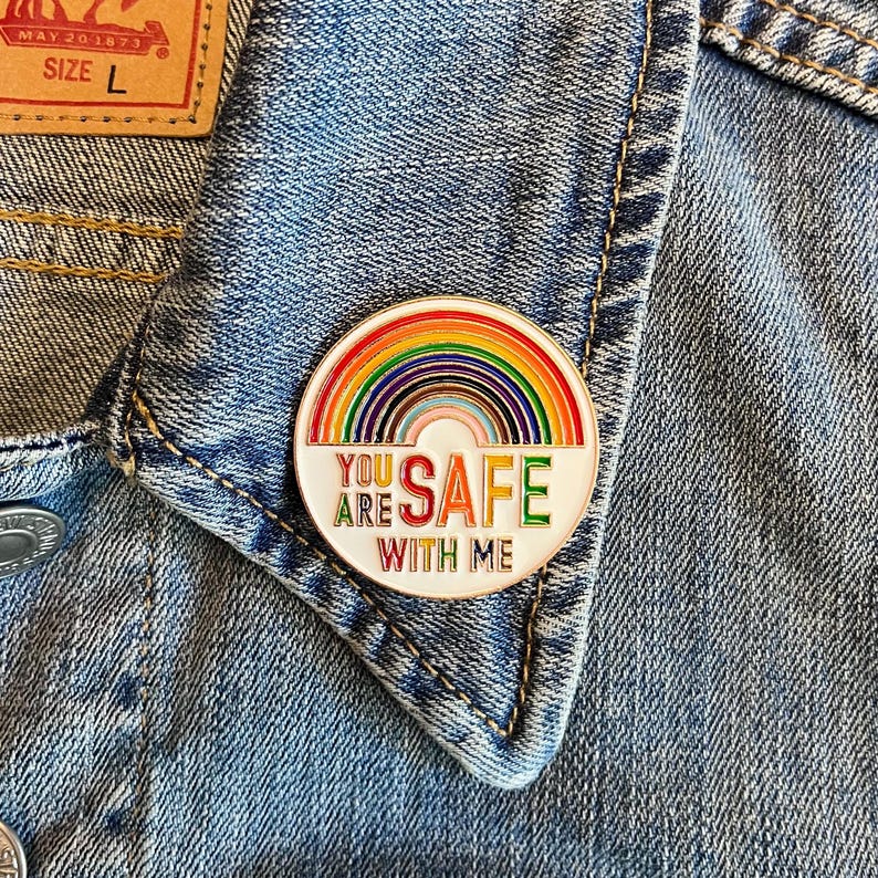 Pu&ograve; includere: Spilla rotonda in smalto con un design arcobaleno e la scritta "YOU ARE SAFE WITH ME" in lettere colorate. La spilla &egrave; applicata su una giacca di jeans.
