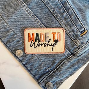 May include: Embroidered patch with the words "MADE TO Worship" in orange, green, and black, with a small black heart. The patch is rectangular with a cream background and a pink border, set against a light blue denim jacket.