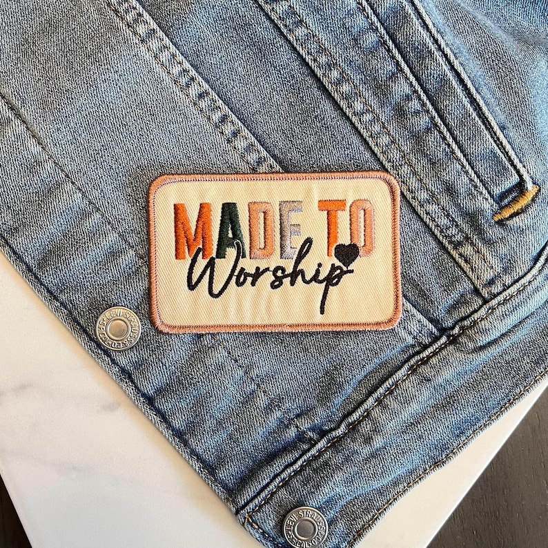 May include: Embroidered patch with the words "MADE TO Worship" in orange, green, and black, with a small black heart. The patch is rectangular with a cream background and a pink border, set against a light blue denim jacket.
