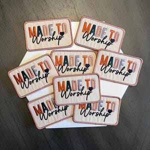 May include: Embroidered rectangular patches with the words "MADE TO Worship" in shades of orange, green, and blue. Each patch has a small black heart. The patches are displayed on a white surface.