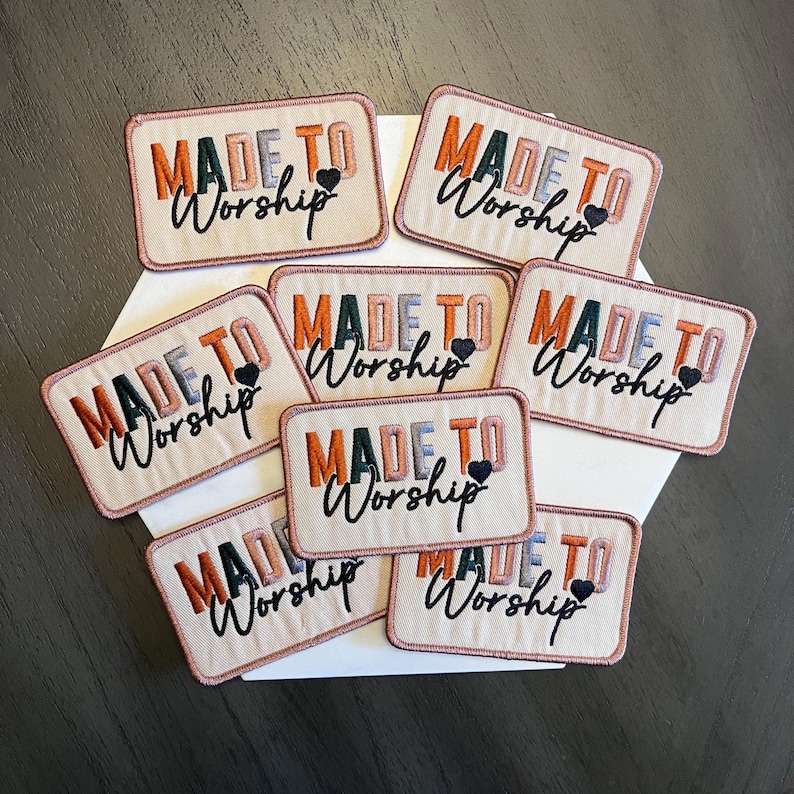 May include: Embroidered rectangular patches with the words "MADE TO Worship" in shades of orange, green, and blue. Each patch has a small black heart. The patches are displayed on a white surface.