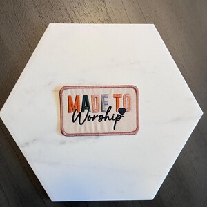 May include: An embroidered patch with the words "MADE TO WORSHIP" in orange, green, grey, and black. A small black heart is next to the word "Worship". The patch is on a white, hexagonal tile.