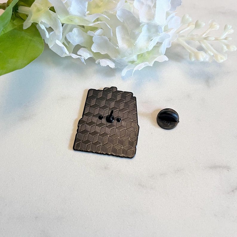 Puede incluir: Un pin de esmalte negro con un patr&oacute;n geom&eacute;trico y un cierre de pin. El pin es rectangular con una parte superior ligeramente curvada. Un cierre de pin negro separado est&aacute; al lado del pin.