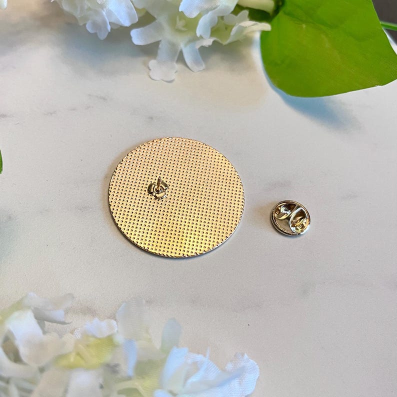 Pu&ograve; includere: Una spilla rotonda color oro con una superficie strutturata e un retro a spillo. La spilla &egrave; accanto a un retro a spillo separato. Lo sfondo &egrave; una superficie chiara con fiori bianchi e foglie verdi.
