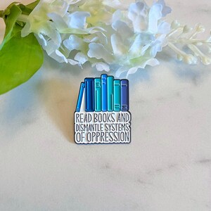 Puede incluir: Pin rectangular de esmalte con una pila de libros en tonos azules y turquesas. El pin tiene el texto "READ BOOKS AND DISMANTLE SYSTEMS OF OPPRESSION" en letras blancas. El pin est&aacute; sobre una superficie blanca y gris.