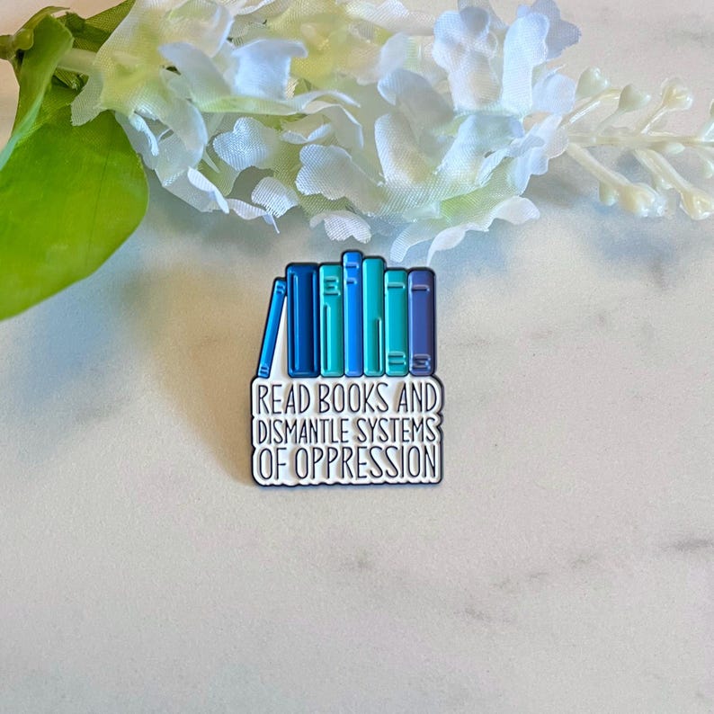 Puede incluir: Pin rectangular de esmalte con una pila de libros en tonos azules y turquesas. El pin tiene el texto "READ BOOKS AND DISMANTLE SYSTEMS OF OPPRESSION" en letras blancas. El pin est&aacute; sobre una superficie blanca y gris.