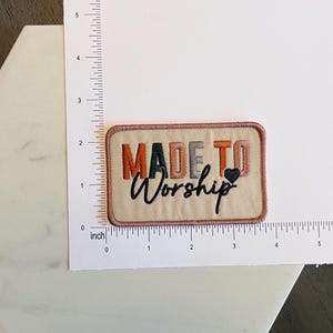 May include: Embroidered rectangular patch with the words "MADE TO Worship" in orange, green, grey, and black. A small black heart is next to "Worship". The patch is approximately 7.6 cm wide.
