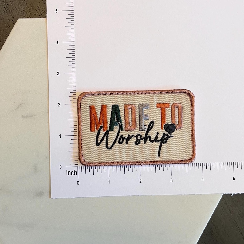 May include: Embroidered rectangular patch with the words "MADE TO Worship" in orange, green, grey, and black. A small black heart is next to "Worship". The patch is approximately 7.6 cm wide.