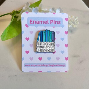 Puede incluir: Un pin de esmalte con una pila de libros azules con el texto "Read Books and Dismantle Systems of Oppression". El pin est&aacute; sobre una tarjeta blanca con corazones rosas y azules y las palabras "Enamel Pins" en la parte superior.