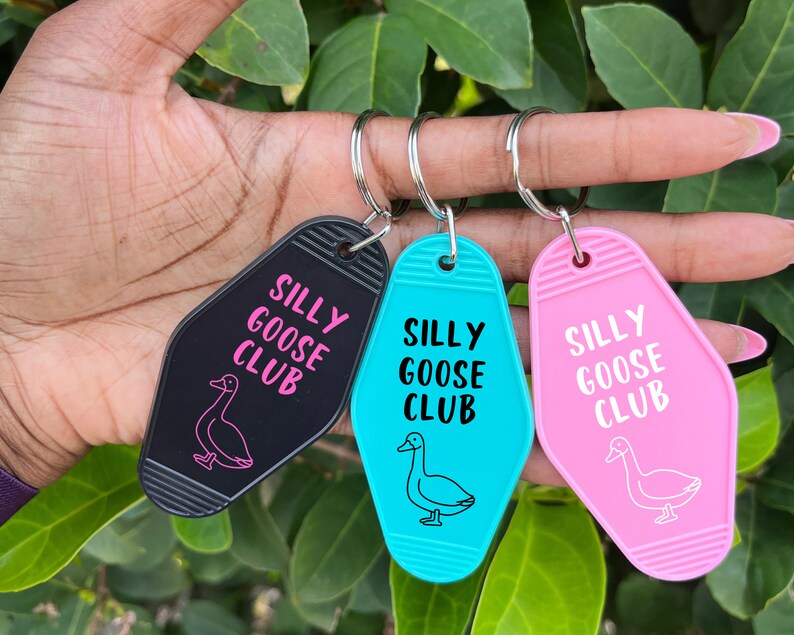 Peut inclure: Trois porte-cl&eacute;s en forme de cl&eacute; de motel. Chaque porte-cl&eacute;s repr&eacute;sente une oie rose avec le texte "Silly Goose Club" en lettres blanches. Les porte-cl&eacute;s sont noirs, turquoise et roses.