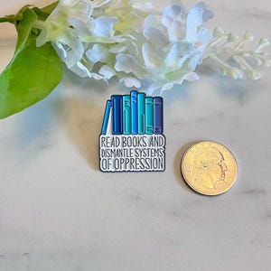 Puede incluir: Pin rectangular de esmalte con una pila de libros en tonos azules y turquesas. El pin tiene el texto "READ BOOKS AND DISMANTLE SYSTEMS OF OPPRESSION" en blanco. Una moneda est&aacute; al lado para la comparaci&oacute;n de tama&ntilde;os.