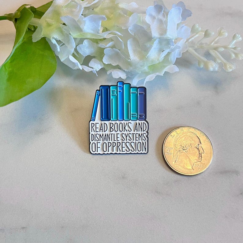 Puede incluir: Pin rectangular de esmalte con una pila de libros en tonos azules y turquesas. El pin tiene el texto "READ BOOKS AND DISMANTLE SYSTEMS OF OPPRESSION" en blanco. Una moneda est&aacute; al lado para la comparaci&oacute;n de tama&ntilde;os.