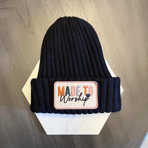 May include: A navy blue ribbed knit beanie hat with a rectangular patch. The patch features the words "MADE TO Worship" in colourful embroidery, with a small heart symbol. The hat is displayed on a white geometric shape.