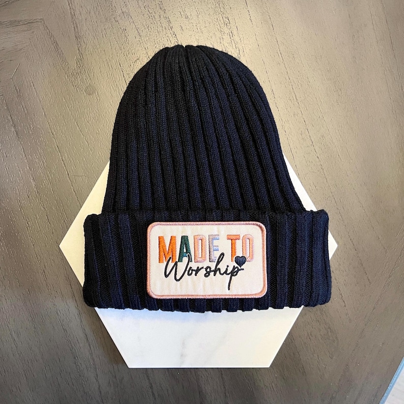 May include: A navy blue ribbed knit beanie hat with a rectangular patch. The patch features the words "MADE TO Worship" in colourful embroidery, with a small heart symbol. The hat is displayed on a white geometric shape.