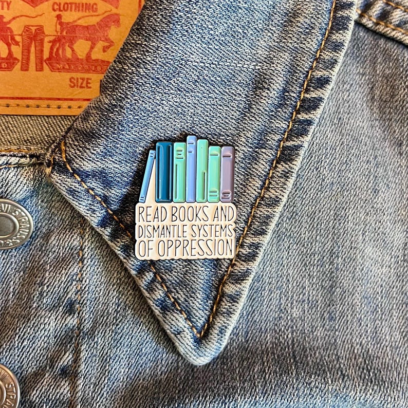 Puede incluir: Un colorido pin de esmalte con una pila de libros en tonos azules y verdes. El pin tiene el texto "READ BOOKS AND DISMANTLE SYSTEMS OF OPPRESSION" en letras blancas. El pin est&aacute; sujeto a una chaqueta vaquera.