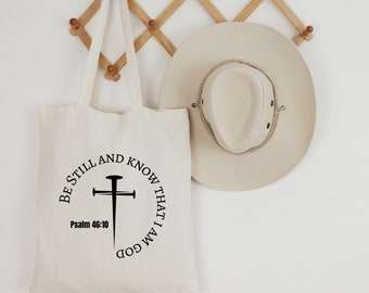 Può includere: Una borsa tote in tela bianca con un design nero che dice "Be still and know that I am God" con una croce e il testo "Psalm 46:10".