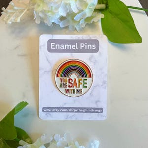 Pu&ograve; includere: Una spilla smaltata colorata con un design arcobaleno e la scritta "YOU ARE SAFE WITH ME". La spilla &egrave; posta su uno sfondo bianco con la scritta "Enamel Pins" in alto. L'URL del negozio Etsy &egrave; visibile in basso.