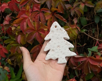 Könnte beinhalten: Ein weißes, flaches, baumförmiges Ornament aus Ton oder Keramik. Die Oberfläche weist ein strukturiertes Muster aus kleinen, geprägten Kreisen und sternartigen Formen auf. Ein kleines kreisförmiges Loch befindet sich oben zum Aufhängen.