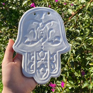 Könnte beinhalten: Eine weiße Keramik-Hamsa-Hand mit aufwendigen geprägten Designs. Der handförmige Wandschmuck weist ein detailliertes Muster aus Wirbeln und geometrischen Formen auf. Sie hat zwei kleine Löcher oben zum Aufhängen, vor dem Hintergrund von grünem Laub und rosa Blüten.